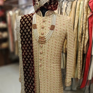 May include: A gold and red embroidered sherwani with a maroon velvet shawl and a pearl and red bead necklace. The sherwani has a high collar and a button closure. The shawl has a gold embroidered border. The necklace is layered and has a decorative pendant.