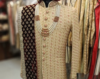 Könnte beinhalten: Ein gold- und rot bestickter Sherwani mit einem maronenfarbenen Samt-Schal und einer Perlen- und roten Perlenkette. Der Sherwani hat einen hohen Kragen und einen Knopfverschluss. Der Schal hat einen goldenen bestickten Rand. Die Kette ist mehrlagig und hat einen dekorativen Anhänger.