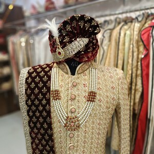 May include: A cream-coloured embroidered men's sherwani with a maroon velvet shawl and a maroon turban with gold embroidery. The sherwani has a pearl and red stone necklace.
