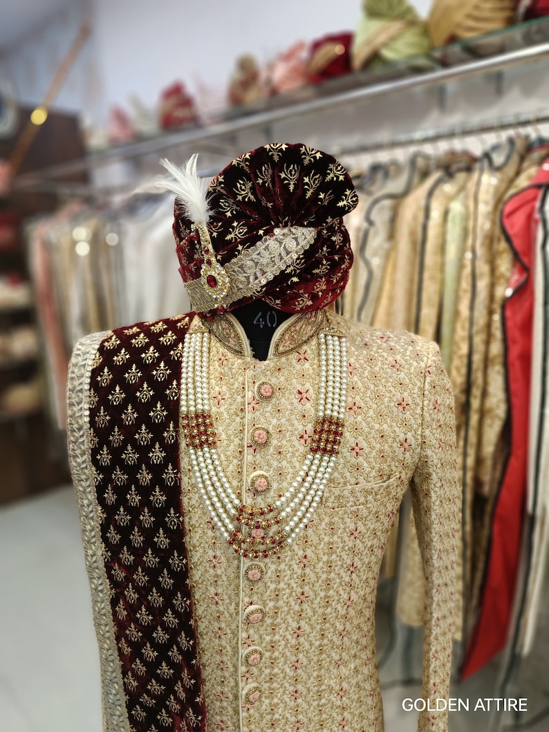 May include: A cream-coloured embroidered men's sherwani with a maroon velvet shawl and a maroon turban with gold embroidery. The sherwani has a pearl and red stone necklace.