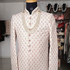 May include: Cream-colored, long-sleeved sherwani with intricate embroidered detailing and red accents. The sherwani features a mandarin collar, decorative buttons, and is adorned with a layered pearl necklace. The garment is displayed on a mannequin.