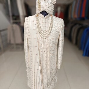 Puede incluir: Un sherwani blanco para hombre bordado con un turbante blanco a juego y un collar de perlas. El sherwani tiene un dise&ntilde;o dorado y plateado y el turbante tiene una pluma en la parte superior. El texto "40" es visible en el sherwani. La imagen est&aacute; etiquetada como "GOLDEN ATTIRE".