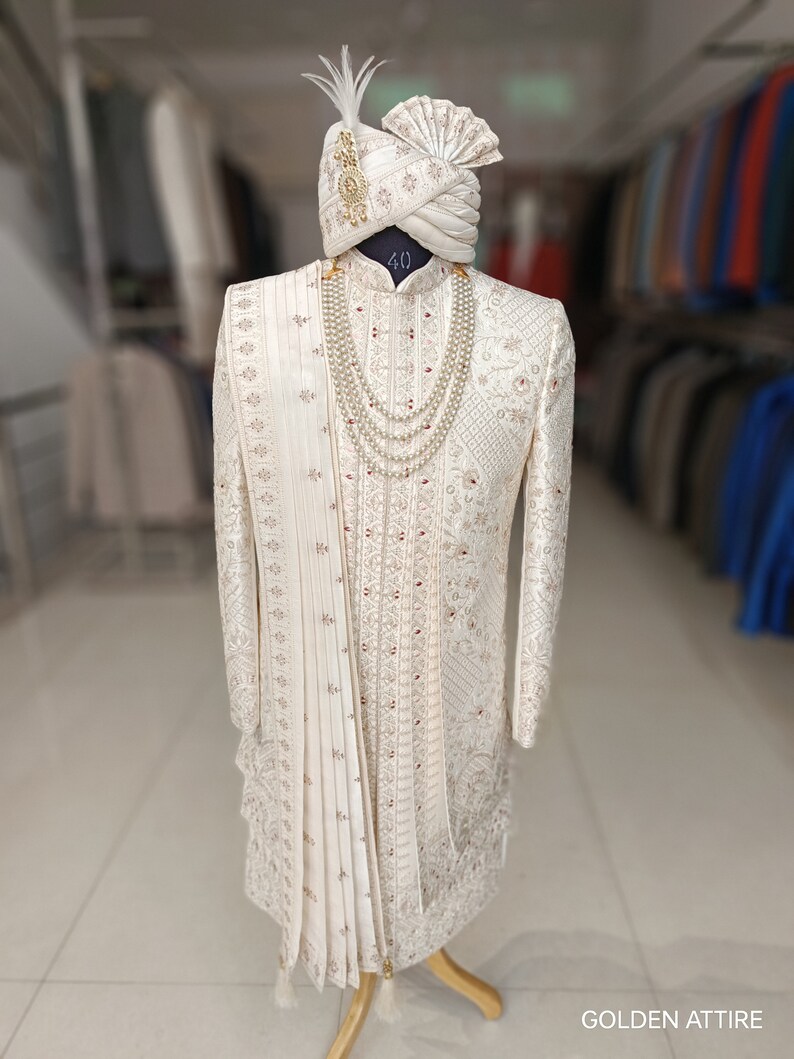 Puede incluir: Un sherwani blanco para hombre bordado con un turbante blanco a juego y un collar de perlas. El sherwani tiene un dise&ntilde;o dorado y plateado y el turbante tiene una pluma en la parte superior. El texto "40" es visible en el sherwani. La imagen est&aacute; etiquetada como "GOLDEN ATTIRE".