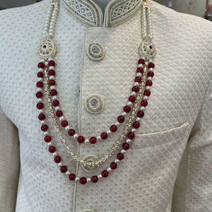 Op de afbeelding: Een witte en gouden kralenketting met rode kralen. De ketting heeft een decoratief hanger met gouden en witte stenen.