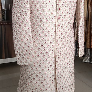 May include: Ivory-colored embroidered sherwani with a mandarin collar, featuring a detailed pattern of red and gold floral motifs. A pearl necklace adorns the neckline. The garment is displayed on a mannequin.