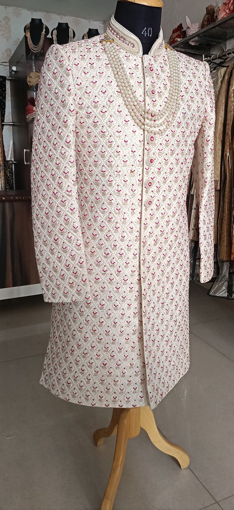 May include: Ivory-colored embroidered sherwani with a mandarin collar, featuring a detailed pattern of red and gold floral motifs. A pearl necklace adorns the neckline. The garment is displayed on a mannequin.