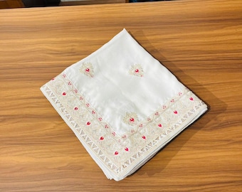 May include: A folded white textile with intricate embroidery. The embroidery features a delicate pattern of cream-colored thread with red accents. The textile is likely a decorative item, such as a scarf or a table runner.
