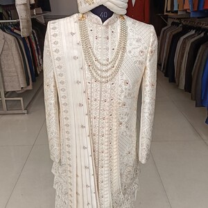 Puede incluir: Un sherwani blanco bordado para hombre con un turbante blanco a juego y un collar de perlas. El sherwani tiene un ribete dorado y una larga estola fluida. El turbante est&aacute; decorado con una pluma. El sherwani se muestra en un soporte de madera. El texto "GOLDEN ATTIRE" es visible en la parte inferior de la imagen.