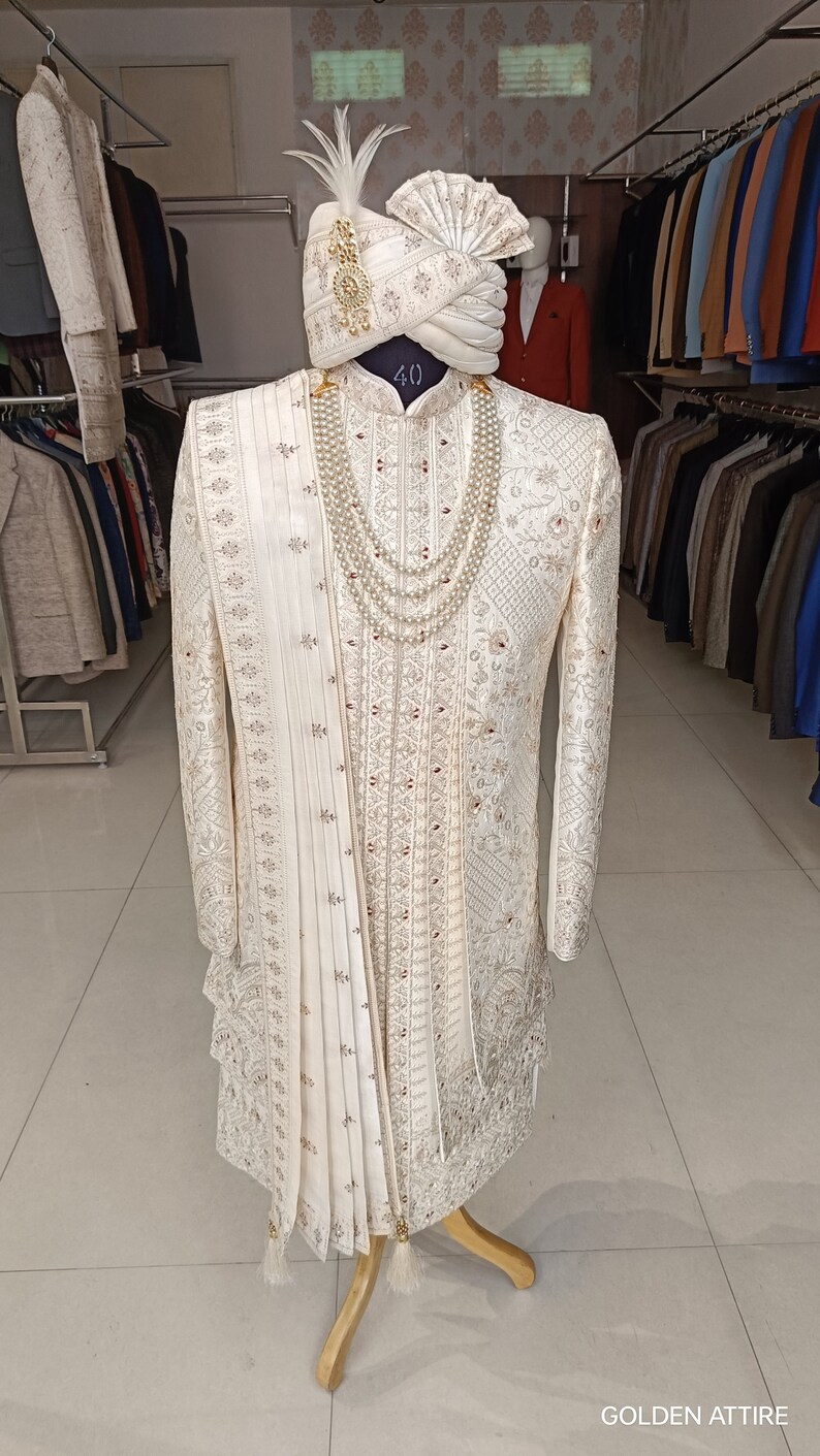 Puede incluir: Un sherwani blanco bordado para hombre con un turbante blanco a juego y un collar de perlas. El sherwani tiene un ribete dorado y una larga estola fluida. El turbante est&aacute; decorado con una pluma. El sherwani se muestra en un soporte de madera. El texto "GOLDEN ATTIRE" es visible en la parte inferior de la imagen.
