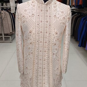 Puede incluir: Una chaqueta blanca bordada para hombre con cuello alto y intrincados patrones florales. La chaqueta est&aacute; adornada con adornos dorados y rojos. La chaqueta se exhibe en un maniqu&iacute; en una tienda de ropa.