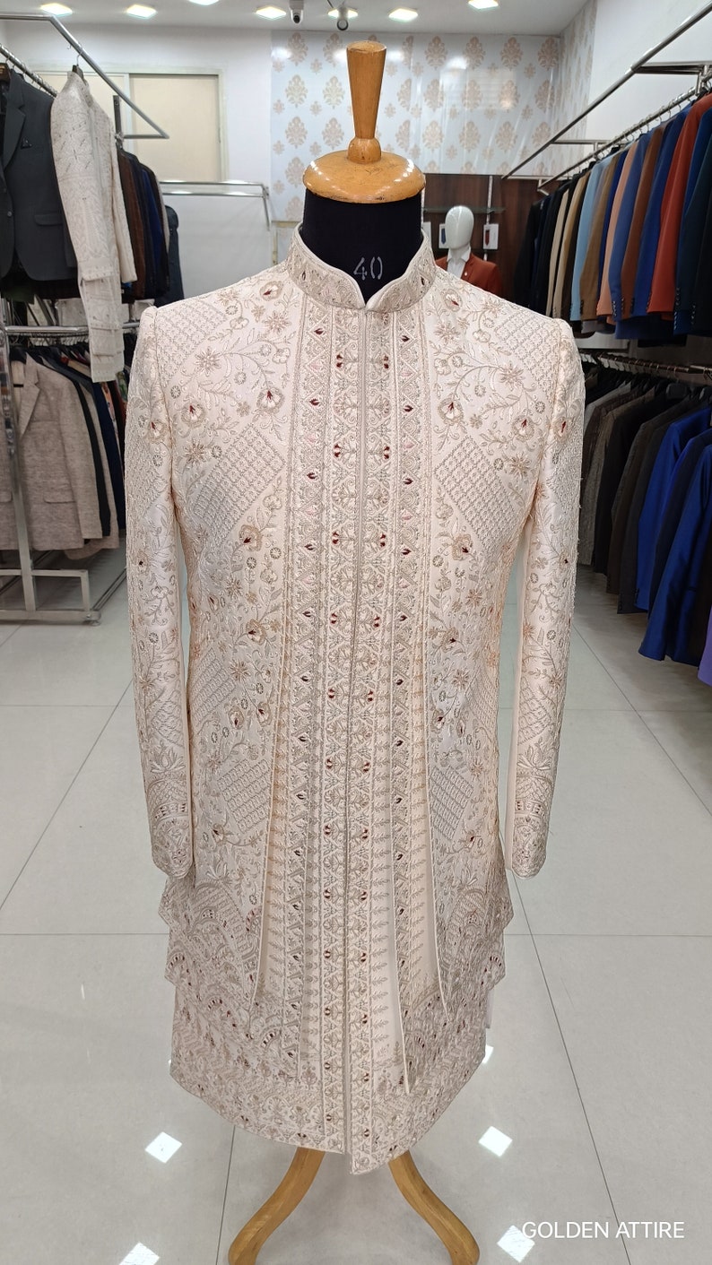 Puede incluir: Una chaqueta blanca bordada para hombre con cuello alto y intrincados patrones florales. La chaqueta est&aacute; adornada con adornos dorados y rojos. La chaqueta se exhibe en un maniqu&iacute; en una tienda de ropa.