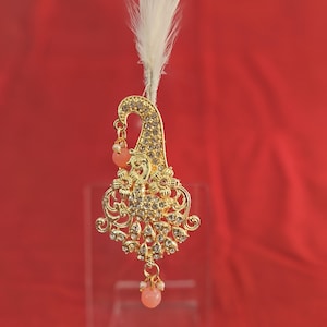 Könnte beinhalten: Ein goldener Haarschmuck in Form eines Pfaus mit weißen Federn und rosa Perlen. Der Schmuck ist mit klaren Strasssteinen verziert.