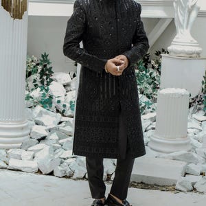K&ouml;nnte beinhalten: Eine schwarze, lang&auml;rmelige, bestickte Jacke mit einem Mandarinkragen und einem langen, flie&szlig;enden Design. Die Jacke wird &uuml;ber einer schwarzen Hose und schwarzen Loafers getragen.