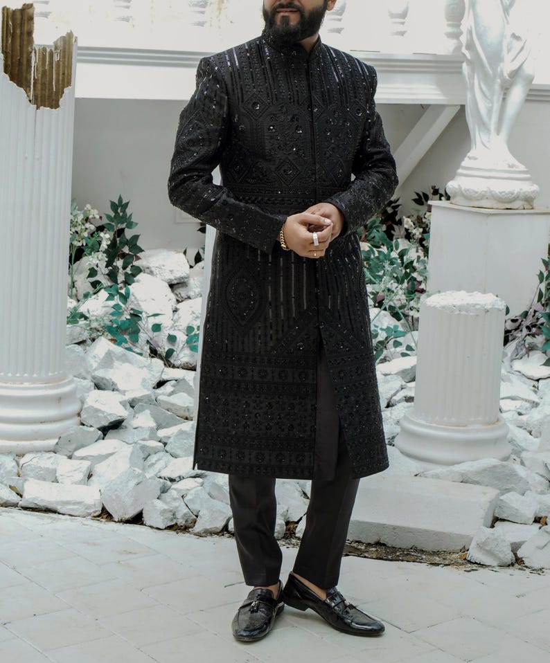 K&ouml;nnte beinhalten: Eine schwarze, lang&auml;rmelige, bestickte Jacke mit einem Mandarinkragen und einem langen, flie&szlig;enden Design. Die Jacke wird &uuml;ber einer schwarzen Hose und schwarzen Loafers getragen.