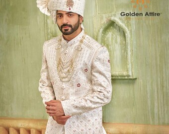 Puede incluir: Un sherwani blanco bordado para hombre con un collar de oro y perlas. El sherwani tiene un cuello alto y un diseño largo y fluido. El hombre lleva un turbante blanco con una joya de oro y roja en la parte superior. El fondo es una pared verde con un diseño dorado y blanco.