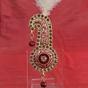 Op de afbeelding: Een versierd decoratief stuk in goud en rood met een witte veer bovenop. Het stuk heeft een gebogen vorm met een ronde onderkant en is versierd met kleine strass steentjes.