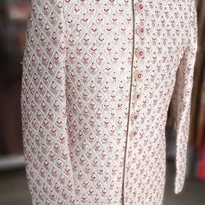 May include: Ivory-colored embroidered jacket with a mandarin collar. The jacket features a diamond pattern with red and gold accents. The garment is displayed on a mannequin with a wooden stand.