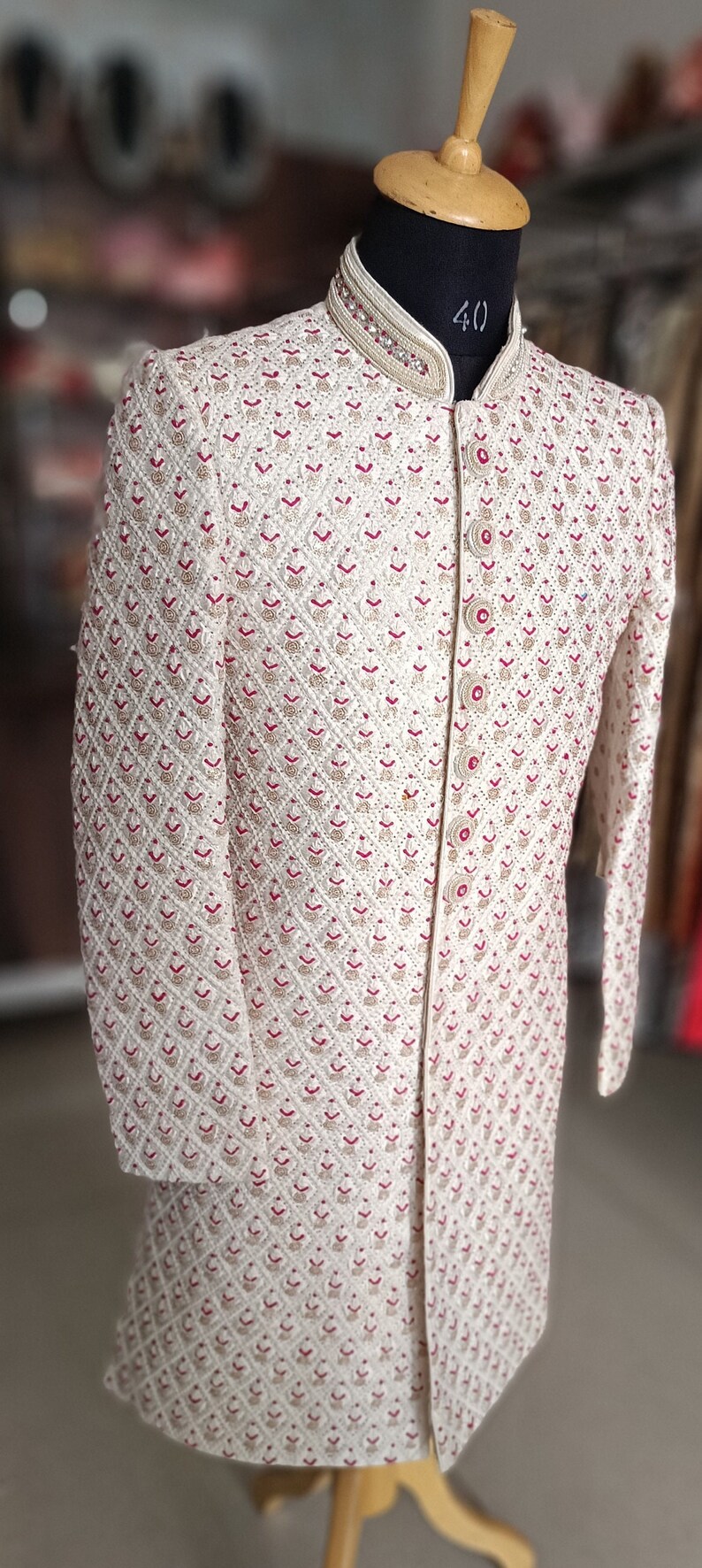 May include: Ivory-colored embroidered jacket with a mandarin collar. The jacket features a diamond pattern with red and gold accents. The garment is displayed on a mannequin with a wooden stand.