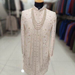 Puede incluir: Un sherwani blanco para hombre bordado con cuello alto y collar de perlas. El sherwani est&aacute; decorado con intrincados patrones florales y acentos rojos. La tela es una mezcla de seda fina. El sherwani se muestra en un maniqu&iacute; en un entorno de tienda.