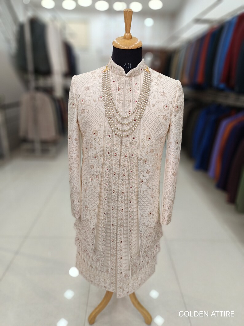 Puede incluir: Un sherwani blanco para hombre bordado con cuello alto y collar de perlas. El sherwani est&aacute; decorado con intrincados patrones florales y acentos rojos. La tela es una mezcla de seda fina. El sherwani se muestra en un maniqu&iacute; en un entorno de tienda.