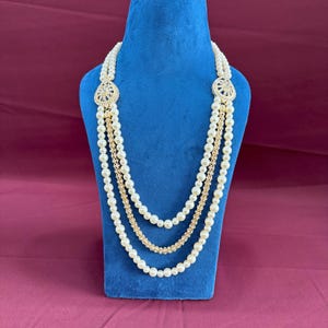 K&ouml;nnte beinhalten: Eine mehrreihige Perlenkette mit goldenen Akzenten und dekorativen Verzierungen. Die Kette wird auf einem blauen Samtst&auml;nder pr&auml;sentiert.