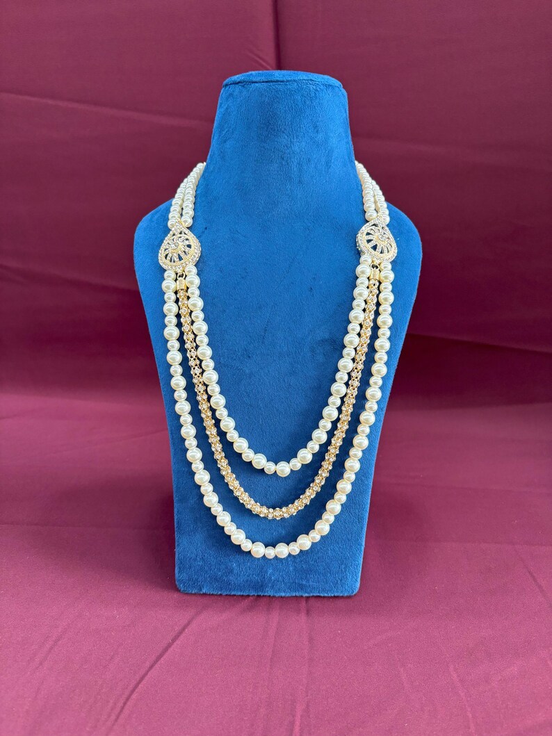 K&ouml;nnte beinhalten: Eine mehrreihige Perlenkette mit goldenen Akzenten und dekorativen Verzierungen. Die Kette wird auf einem blauen Samtst&auml;nder pr&auml;sentiert.