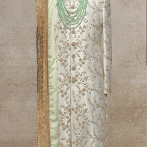 K&ouml;nnte beinhalten: Ein wei&szlig;er Sherwani mit Blumenmuster und ein hellgr&uuml;ner Schal mit goldenen Verzierungen. Der Sherwani hat einen hohen Kragen und einen Knopfverschluss. Eine hellgr&uuml;ne Perlenkette wird &uuml;ber dem Sherwani getragen.
