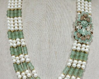Puede incluir: Un collar de varias hebras con perlas blancas y cuentas verde claro. El collar tiene un colgante floral decorativo con detalles en verde y marrón. El collar se exhibe en un maniquí.