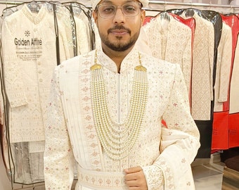 May include: A man wearing a white sherwani with gold embroidery and a white turban with gold accents. He is wearing a pearl necklace and a white sash. The man is standing in front of a rack of clothing.