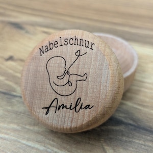 Könnte beinhalten: Eine Holzkiste mit Deckel, graviert mit den Worten "Nabelschnur" und "Amelia". Die Kiste zeigt eine einfache Strichzeichnung eines Babys im Mutterleib mit Nabelschnur.