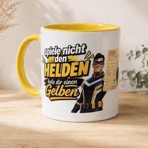 Könnte beinhalten: Eine weiße Keramiktasse mit gelbem Innenraum und Henkel, bedruckt mit einem Superhelden im blau-gelben Anzug. Der Text lautet "spiele nicht den HELDEN hole dir einen Gelben" neben einem kleinen "Arbeitsunfähigkeitsbescheinigung"-Formular.