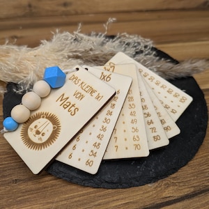 Könnte beinhalten: Multiplikationskarten aus Holz mit einem Löwenmotiv und dem Text "DAS KLEINE 1x1 VON Mats". Die Karten sind an einer Holzkette mit blauen Akzenten befestigt. Die Karten sind fächerförmig angeordnet.