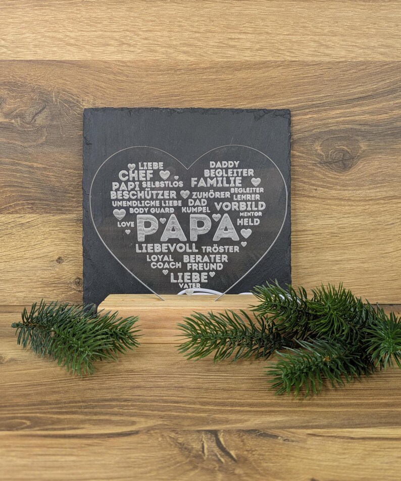 Op de afbeelding: Een zwarte leien hartvormige plaat met het woord "PAPA" in het midden, omringd door woorden als "LOVE", "DAD" en "FAMILY". De plaat staat op een houten voet, met dennentakken aan weerszijden. De achtergrond is een houten oppervlak.
