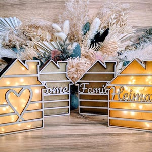 Könnte beinhalten: Vier holzförmige LED-Leuchten in Hausform mit warmweißer Beleuchtung. Eine hat einen Herz-Ausschnitt, andere die Wörter "home", "family" und "Heimat". Die Leuchten stehen auf einer Holzoberfläche.