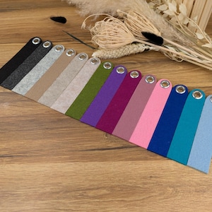 Op de afbeelding: Een verzameling gekleurde viltstroken met metalen oogjes, diagonaal geplaatst op een houten oppervlak. De stroken variëren in kleur, waaronder zwart, grijs, beige, groen, paars, roze, blauw en turquoise. Droogbloemen en een houten kistje staan op de achtergrond.