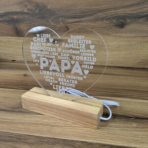 Op de afbeelding: Een helder acryl hartvormig licht met het woord "PAPA" in grote letters. Het hart is ge&euml;tst met woorden als "LIEFDE", "VADER" en "FAMILIE". Het staat op een houten voet met een wit snoer.