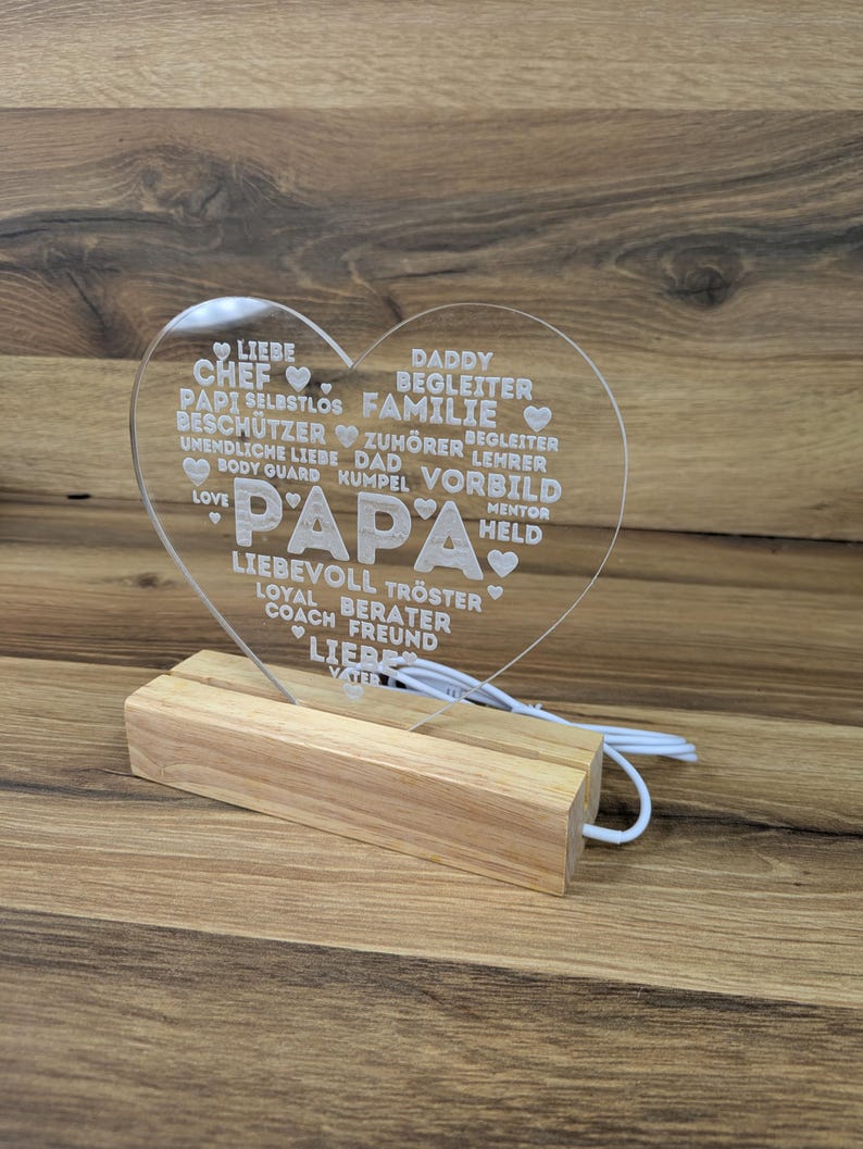 Op de afbeelding: Een helder acryl hartvormig licht met het woord "PAPA" in grote letters. Het hart is ge&euml;tst met woorden als "LIEFDE", "VADER" en "FAMILIE". Het staat op een houten voet met een wit snoer.