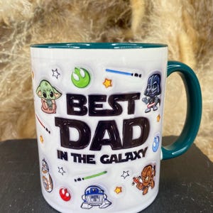 Op de afbeelding: Witte keramische mok met een teal handvat en rand, versierd met Star Wars-personages en de tekst "BEST DAD IN THE GALAXY". De mok heeft afbeeldingen van Baby Yoda, Darth Vader en andere iconische personages.