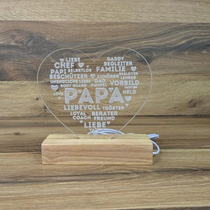 Op de afbeelding: Een hartvormig, helder acryl LED-nachtlampje met het woord "PAPA" in grote letters. Het hart is gevuld met woorden als "LIEFDE", "VADER" en "FAMILIE". Het staat op een rechthoekige houten basis.