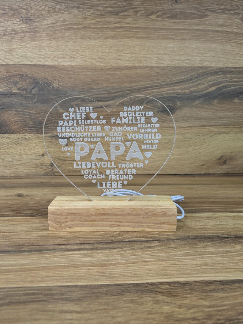 Op de afbeelding: Een hartvormig, helder acryl LED-nachtlampje met het woord "PAPA" in grote letters. Het hart is gevuld met woorden als "LIEFDE", "VADER" en "FAMILIE". Het staat op een rechthoekige houten basis.