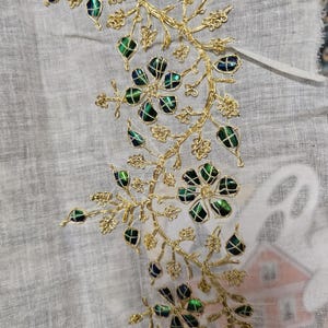 May include: Close-up of a textile with gold-colored floral embroidery. The embroidery features intricate designs with green and blue iridescent accents, creating a shimmering effect. The fabric is a light, neutral color.