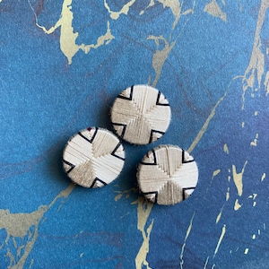 Puede incluir: Tres botones blancos con diseños tejidos en negro y rojo. Los botones están sobre una superficie de mármol azul y dorado.