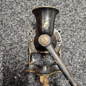 Puede incluir: Un antiguo molinillo de café negro con un mango de madera. El molinillo tiene una parte superior en forma de embudo y una manivela. Las palabras "SPONG & CO" y "ENGLAND" son visibles en el cuerpo del molinillo. El molinillo está montado sobre una superficie oscura.