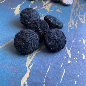 Puede incluir: Cinco botones negros con textura tejida. Los botones están dispuestos en un grupo sobre una superficie de mármol azul y dorado.