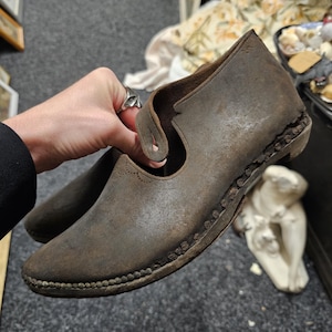 May include: A pair of antique brown leather shoes with a strap and decorative stitching. The shoes have a worn appearance, suggesting age and use. The sole is detailed with small, rounded accents.