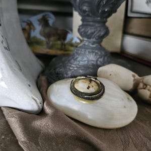 May include: An antique, oval-shaped trinket box in white, with a silver-coloured metal ring around the lid. The box rests on a brown fabric surface, with a decorative metal candlestick and other vintage items in the background.