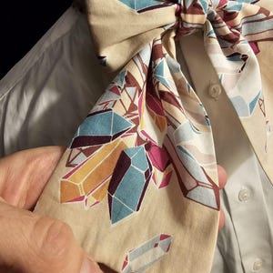 May include: A beige bow tie with a geometric pattern of colorful crystals. The bow tie is tied around a white shirt collar.