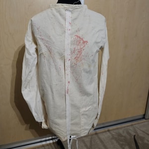 Könnte beinhalten: Cremefarbene Zwangsjacke mit langen Ärmeln, ausgestellt auf einer schwarzen Schaufensterpuppe. Die Jacke hat einen vertikalen weißen Streifen auf dem Rücken und ist mit roten Flecken bespritzt. Die Jacke ist an den Handgelenken und an der Taille gebunden.