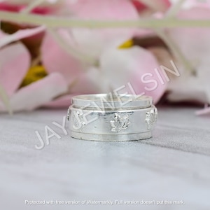 May include: A silver ring with a spinning band featuring a design of maple leaves. The ring is on a light grey surface.