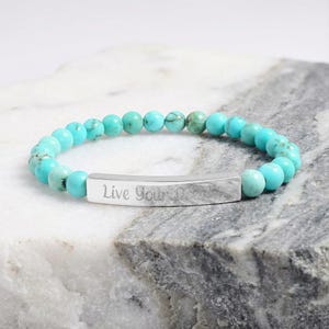 Könnte beinhalten: Ein türkisfarbenes Perlenarmband mit einem silbernen Balken, auf dem die Worte "Live Your Dreams" eingraviert sind. Das Armband besteht aus runden, polierten türkisfarbenen Perlen und einem gebogenen Silberbalken.